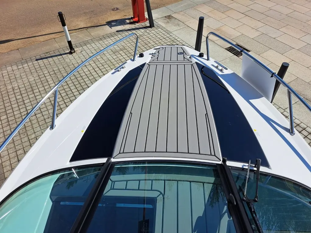 Top view of a boat's deck featuring a gray non-slip mat, metal railing, and glass windshield against a stone pavement background.